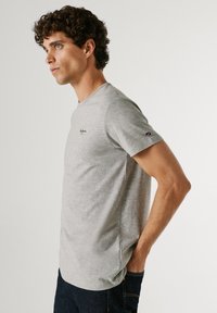 Jonge man met krullend haar die een lichtgrijs T-shirt met korte mouwen en donkere jeans draagt, staand in profiel met één hand in de zak.