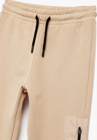 Beige Jogginghose aus weichem Stoff, mit einem elastischen Bund und schwarzen Kordeln sowie einer seitlichen Reißverschlusstasche.