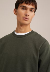 Sudadera verde oliva hecha de tejido texturizado, con cuello redondo y cuello acanalado, llevada sobre una camisa blanca. Vista en primer plano del ajuste y los detalles.