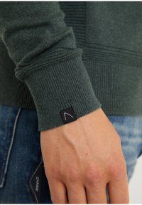 Dunkelgrüner gestrickter Pulloverbund mit Rippenstruktur und einem schwarzen Etikett mit weißem Logo, kombiniert mit blauen Jeans.