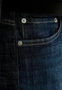 Detailný záber na tmavomodré rifľové vrecko s viditeľnými stehmi a kovovými nitmi na textúre látky.