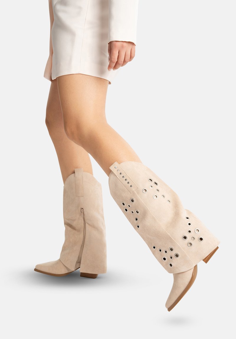 Jambes portant des bottes de cowboy en daim beige avec des œillets en métal, assorties à une mini-jupe beige et un haut coordonné sur un fond uni.