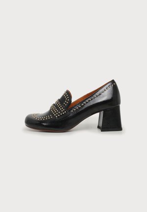 Mocasín de cuero negro con tacón bloque, con detalles de tachuelas doradas a lo largo de los bordes y la correa, con puntera redondeada y forro interior marrón.
