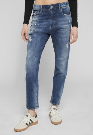 Isik kannab siniseid kulunud sirge säärega teksapükse ja valgeid tossusid mustade triipudega, seisab ühevärvilise halli tausta ees.