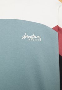 Barevně blokovaná tkanina s bílými, tyrkysovými, rudo-oranžovými a černými sekcemi, s bílým vyšívaným textem "downtown KAOTIKO" na tyrkysové oblasti.