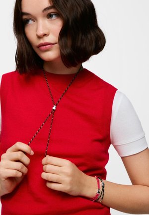 Giovane donna con capelli castani corti che indossa un top rosso senza maniche sopra una camicia bianca a maniche corte, tiene una collana di corda nera e indossa braccialetti abbinati.