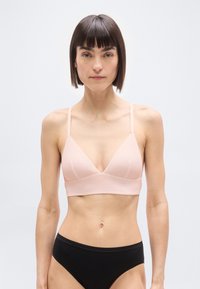 Γυναίκα με κοντά σκούρα μαλλιά που φοράει ροζ ανοιχτό μπραλέτο και μαύρο εσώρουχο, κοιτάζοντας μπροστά σε απλό ανοιχτό φόντο.