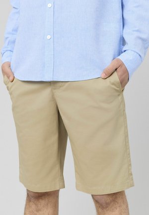 Shorts beiges en tissu léger, avec poches latérales, coupe droite et ourlet cousu, associés à une chemise bleu clair à boutons.