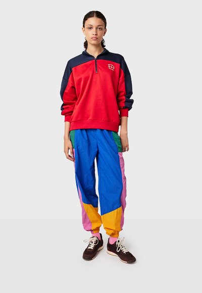 Rode en marineblauwe sweatshirt met rits en lange mouwen, gecombineerd met multicolore, losse trainingsbroek met blauwe, groene, roze en gele secties.