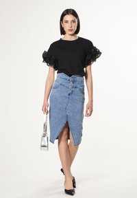 Camiseta negra con mangas de encaje con volantes, combinada con una falda lápiz de mezclilla azul clara que presenta un slit frontal y cierre de cinco botones. Bolso plateado.
