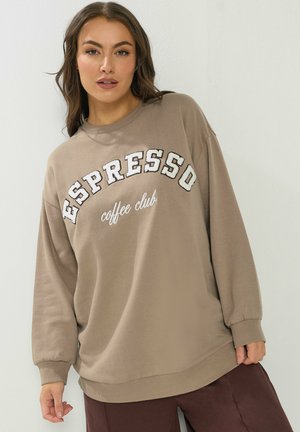 Frau trägt einen beigen, übergroßen Pullover mit dem Text „ESPRESSO coffee club“, kombiniert mit dunkelbrauner Hose, posiert vor einer weißen Wand.