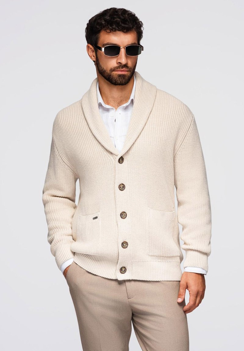 Beige gestrickte Strickjacke mit einem Schalkragen, fünf Knöpfen und zwei vorderen Taschen. Getragen über einem hellen Hemd mit Knöpfen.