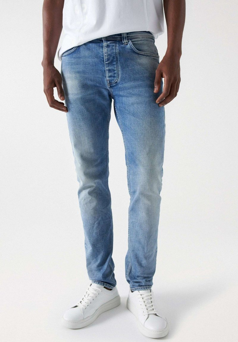Light blue denim jeans with a slim fit and faded texture, featuring five-pocket design and belt loops, paired with white sneakers.