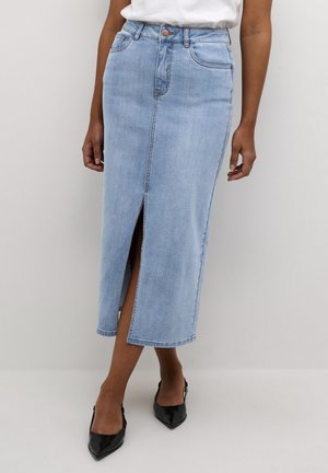 Donna che indossa una gonna midi in denim azzurro chiaro con spacco frontale e scarpe nere con punta affilata su sfondo semplice.
