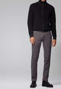 Svart zip-up jacka med hög krage, grå skräddarsydda byxor och svarta dressskor. Slät tygkvalitet med minimalistisk design.