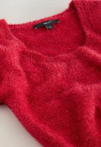 Czerwony sweter z dzianiny o puszystej fakturze, szerokim dekolcie i widoczną metką na szyi. Miękki, lekki materiał o lekko luźnym kroju.
