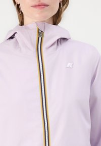 Jachetă de culoare violet deschis, cu glugă și fermoar complet. Are o dungi multicolore de-a lungul fermoarului și un logo mic pe piept.