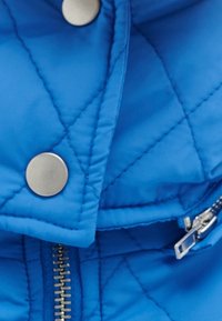 Chaqueta acolchada azul con botones de presión plateados y cremallera. La tela parece suave con patrones de costura ondulados.