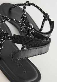 Sandalias negras con tiras trenzadas en blanco y negro, con suela plana y acentos de cuero negro texturizado, diseñadas para ofrecer comodidad y flexibilidad.