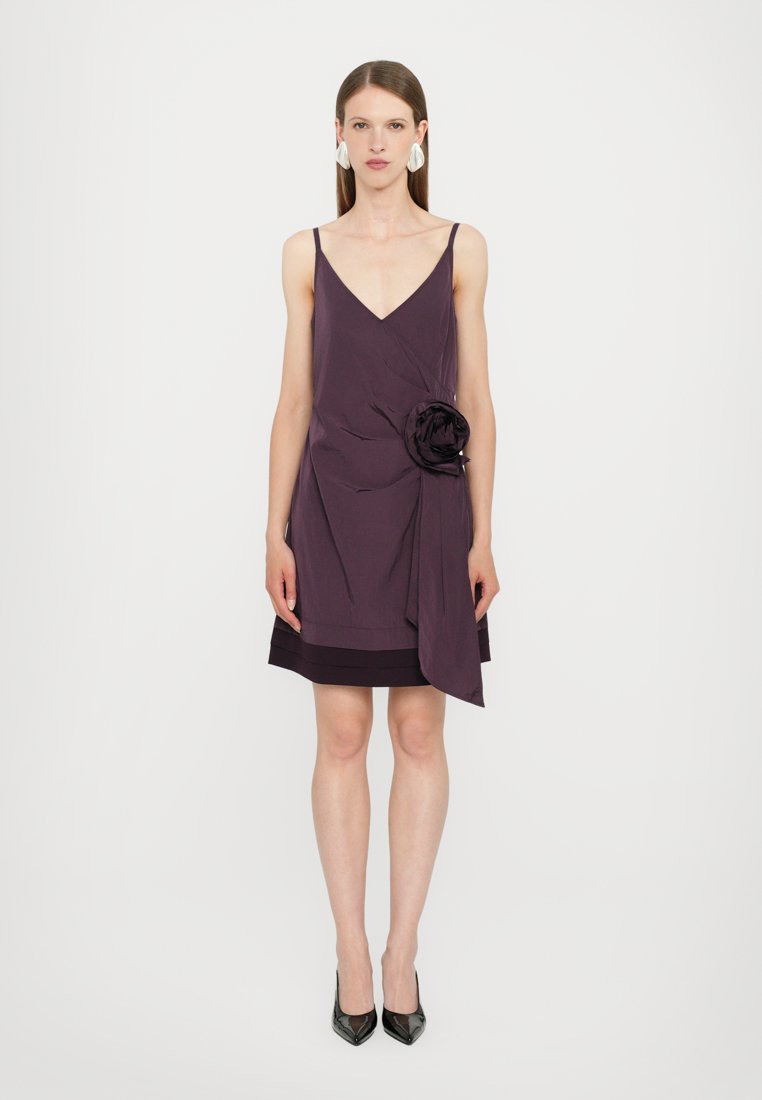 Robe violette avec un décolleté en V, tissu drapé, détail floral sur le côté et ourlet en couches. Texture lisse et coupe ajustée, accompagnée de talons noirs.