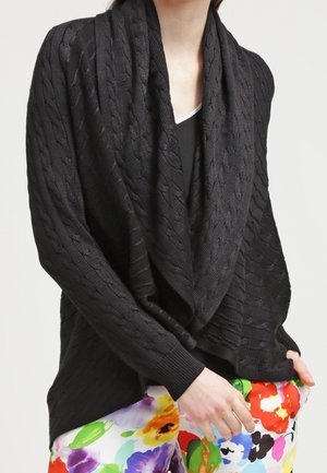 Zwarte kabelgebreide gedrapeerde cardigan gedragen over een zwarte top, gecombineerd met kleurrijke bloemmotiefbroeken, bij een persoon tegen een effen achtergrond.