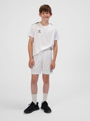 Garçon souriant aux cheveux bruns courts portant une tenue de sport blanche et des baskets noires, debout devant un fond uni gris clair.