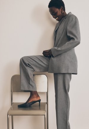 Grå skräddarsydd kostym med texturerad yta, kombinerad med spetsiga svarta mules. Modellen poserar med ett ben på en silverstol, minimalistisk bakgrund.