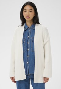 Mladá žena v bielom rebrovanom kardig�áne cez modrú košeľu s gombíkmi a zodpovedajúcimi džínsami na jednoduchom pozadí.