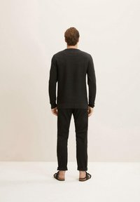 Zwart gestructureerd sweater met lange mouwen, ontspannen pasvorm; gecombineerd met donkere broeken en open-teentjes sandalen. Minimalistische achtergrond, neutrale tinten.