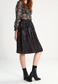 Blouse noire transparente avec imprimé floral, manches longues et poignets volants ; associée à une jupe midi plissée noire et des bottines noires.