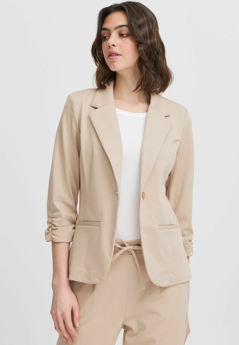 Beige blazer med slagkrage, enknappstängning och två framfickor. Mjuk, texturerad tyg med uppvikta ärmar för en avslappnad passform.