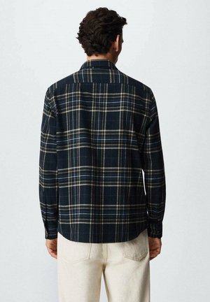 Mann trägt dunkles kariertes Hemd mit Knöpfen und hellbeige Hose, steht mit dem Rücken zur Kamera vor einfachem Hintergrund.