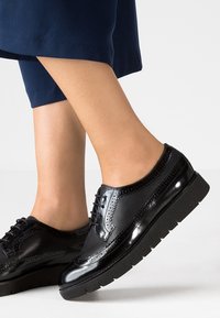 Chaussures en cuir verni noir avec des détails brogue, dotées d'une semelle noire épaisse et de lacets ronds. Texture lisse avec des perforations décoratives.