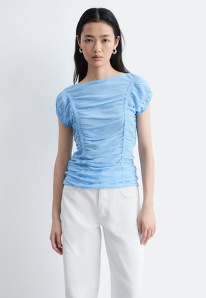 Frau mit glatten schwarzen Haaren, die silberne Creolen, ein gerafftes hellblaues Kurzarmtop und weiße, hoch taillierte Hosen trägt.