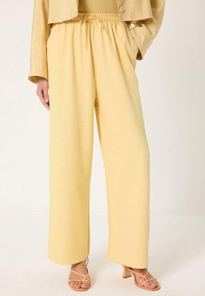 Γυναίκα που φοράει παντελόνι ανοιχτού κίτρινου χρώματος, ψηλόμεσο και με φαρδιά μπατζάκια, με ελαστική μέση και κορδόνι, συνδυασμένο με σανδάλια με λουράκια και τακούνι σε γυμνό χρώμα.