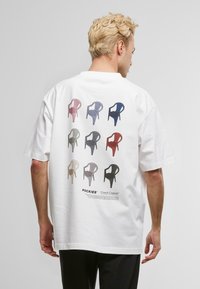 Hvit t-skjorte med grafisk trykk av fargerike plaststoler i forskjellige nyanser, arrangert i to rader på baksiden.