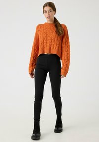 Mujer con cabello castaño recogido en una coleta baja, vestida con un suéter corto de punto trenzado color naranja, leggings negros y botines negros, de pie sobre un fondo blanco.