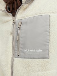 Chaqueta de forro polar color crema con acentos grises, presenta un cierre con cremallera en la parte frontal, un bolsillo con parche y logo, y detalle de tejido texturizado.