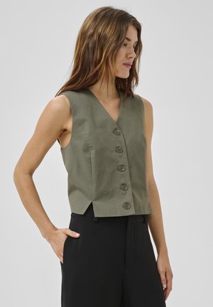 Kobieta ubrana w bezrękawnik w kolorze oliwkowym z guzikami i czarne spodnie, stojąca z jedną ręką w kieszeni, patrząca w prawo.