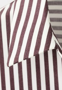 Primer plano del cuello de una camisa a rayas verticales marrones y blancas, con punta en forma de pico y costuras visibles.