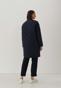 Vrouw in een lange gewatteerde marineblauwe jas met diamantstiksels, zwarte cropped broek en witte sneakers, staand tegen een effen lichte achtergrond.