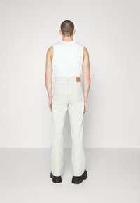 Pantalones de mezclilla de corte ancho y color claro con un parche de cuero marrón en la cinturilla trasera; combinados con una blusa sin mangas blanca.
