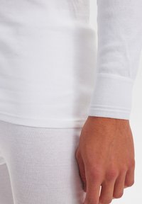 White long-sleeve shirt with a ribbed texture. Features a fitted design and a snug cuff at the wrist, paired with white fitted bottoms.