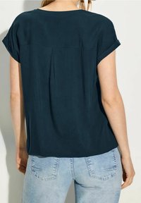 Donna che indossa una blusa corta a maniche corte di un blu scuro, con una piega sul retro, abbinata a jeans in denim azul chiaro, mostrata di spalle.