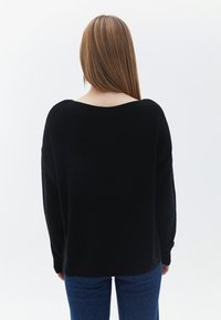 Černý pletený svetr s širokým výstřihem a spadlými rameny, zobrazený ze zadní strany. V kombinaci s modrými džíny.