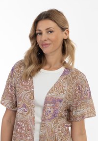Kimono léger avec motifs à manches courtes, dans un design paisley marron, crème et violet, porté sur un haut ajusté blanc.