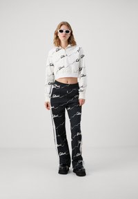 Sudadera blanca cortada con un patrón de escritura negra y cremallera. Pantalones negros con un patrón similar, franjas laterales y un corte relajado. Zapatillas negras de plataforma.
