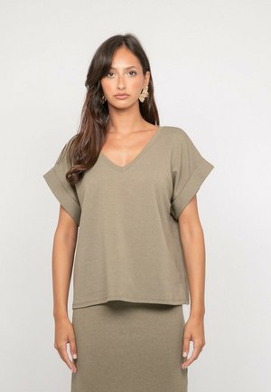 Femme aux longs cheveux bruns portant un haut beige à manches courtes en V et une jupe assortie, debout devant un fond clair uni.