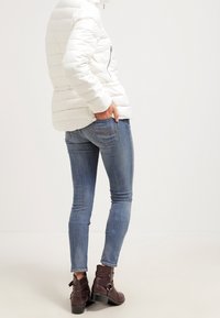 Bílá puffová bunda s horizontálním prošíváním a boční kapsou na zip, kombinovaná s modrými skinny džíny a hnědými kotníkovými botami se sponami.