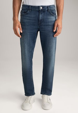 Mężczyzna ubrany w ciemnoniebieskie dopasowane dżinsy i białe trampki, stojący z rozluźnionymi rękami wzdłuż ciała na neutralnym tle.